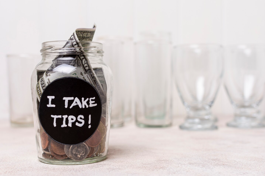Glass tip jar labeled ‘I take tips!’, illustrating modern tipping etiquette